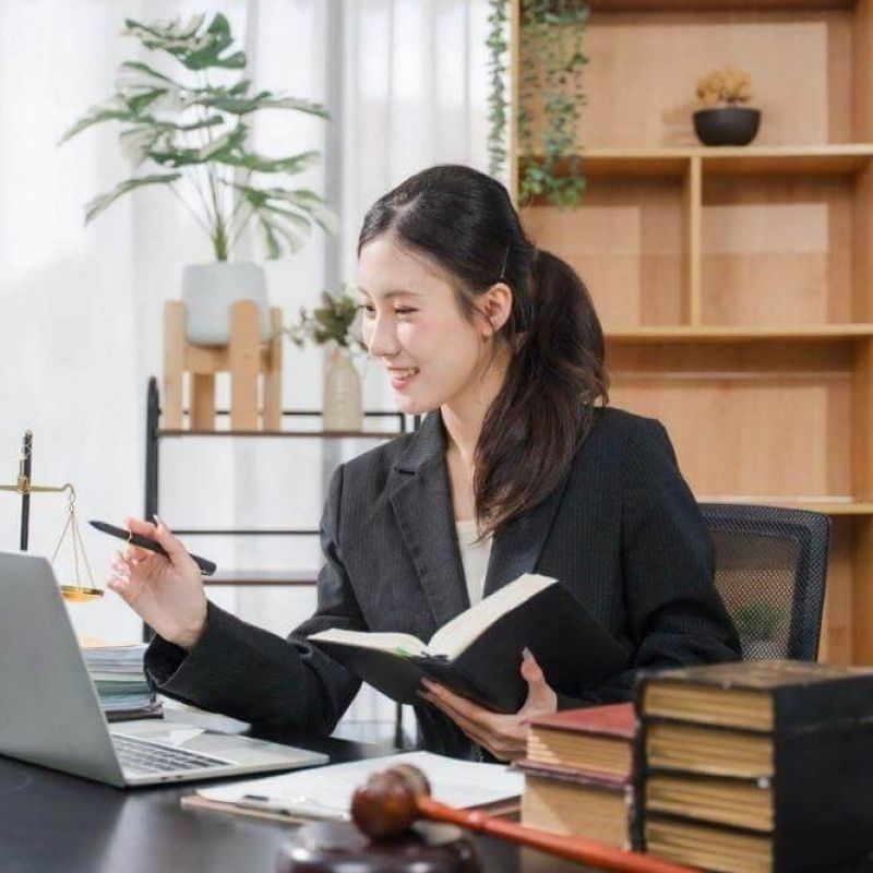 Professional woman in a blazer sits at a desk with a laptop, an open notebook, a gavel, and stacked books in a tidy office.