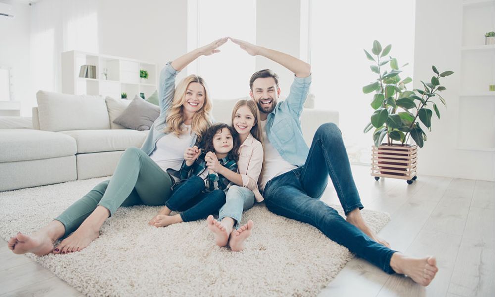 Happy family sitting on carpet in bright living room, smiling and forming a roof shape with their arms.