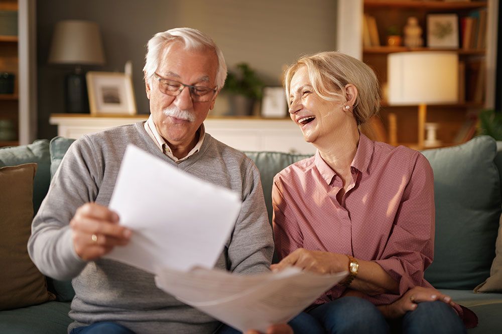 Elderly couple happily reviewing documents on a sofa at home, smiling and enjoying each other's company.