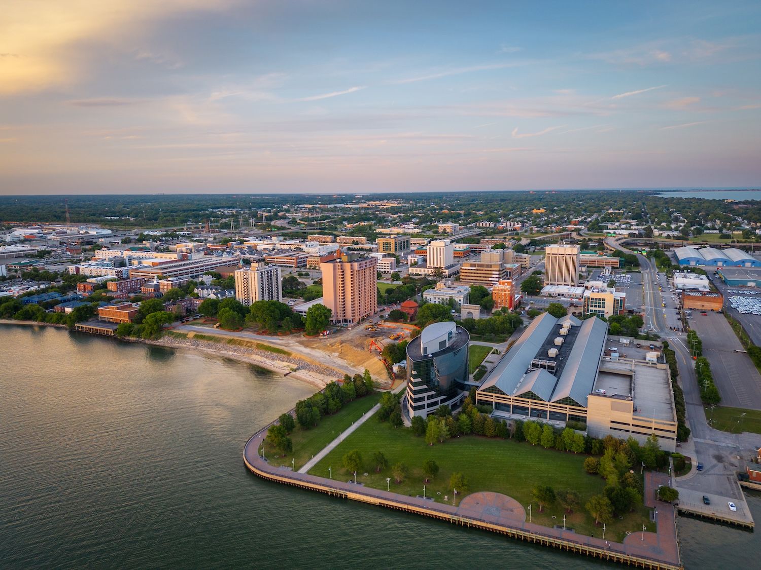 Aerial view of waterfront cityscape during sunset, highlighting green spaces and urban architecture by the water.