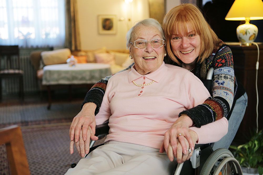 Elderly woman in a wheelchair smiling with a caregiver in a cozy living room setting.
