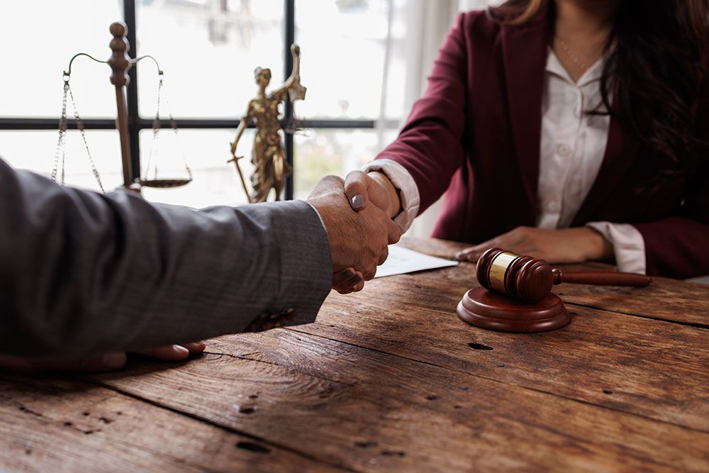 Handshake between two professionals at a wooden desk with gavel, scales, and Lady Justice statue in the background.