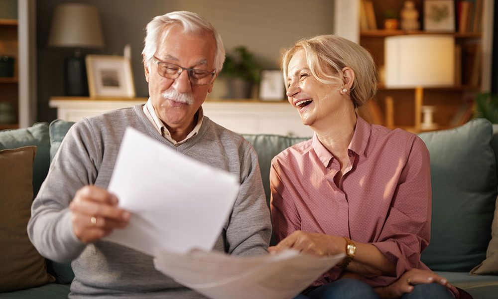 Elderly couple happily reviewing documents on a sofa at home, smiling and enjoying each other's company.