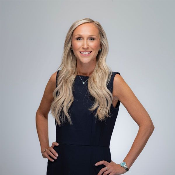 Shannon Laymon-Pecoraro in a navy dress with long blonde hair and her hands on hips, posing confidently against a plain background.