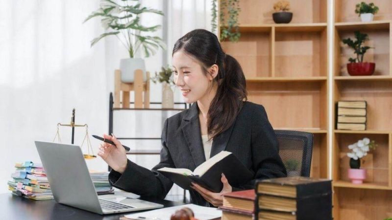 Professional woman in a blazer sits at a desk with a laptop, an open notebook, a gavel, and stacked books in a tidy office.