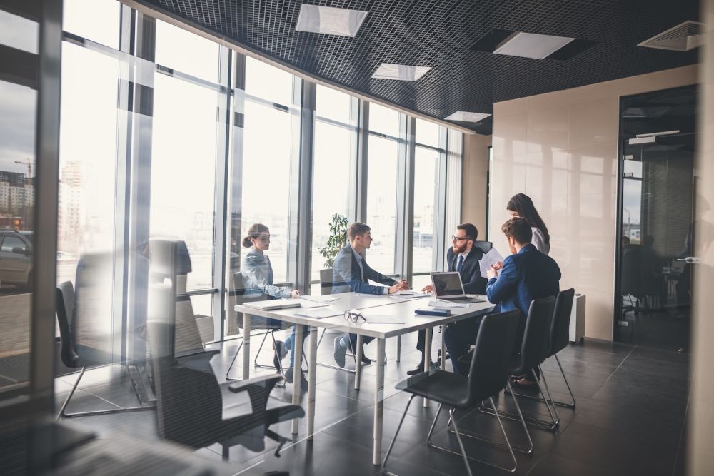 Business meeting in modern office with large windows, five professionals collaborating at a table with laptops.