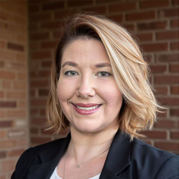Brittany J. Moore with short blonde hair, wearing a black blazer, in front of a brick wall.
