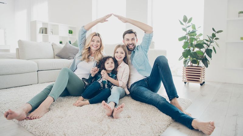 Happy family sitting on carpet in bright living room, smiling and forming a roof shape with their arms.