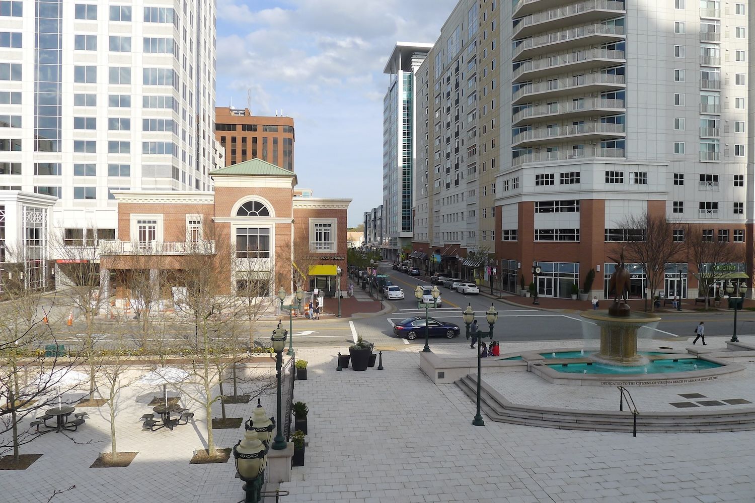 Urban cityscape with tall buildings, street view, and a central fountain on a sunny day.