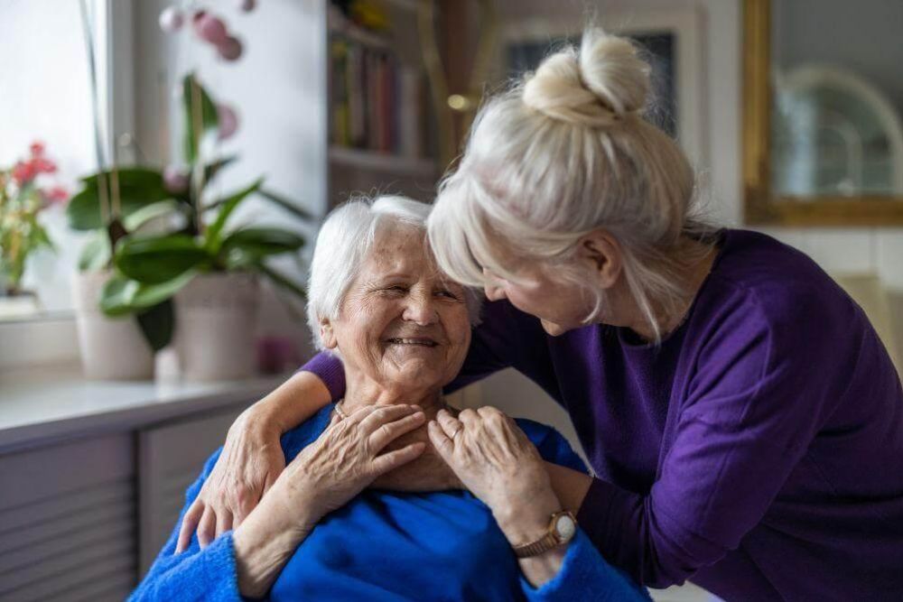 Caregiver in a purple sweater helps an elderly woman in blue with a necklace, both smiling indoors at home.