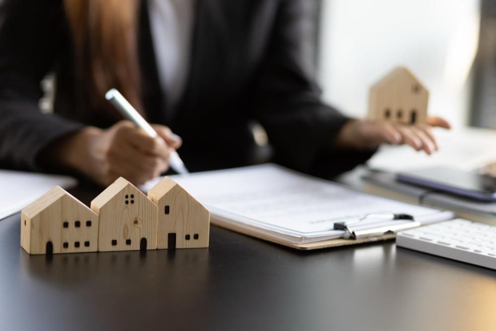 Miniature wooden houses lined up on a desk as a person signs paperwork, suggesting real estate or mortgage planning.