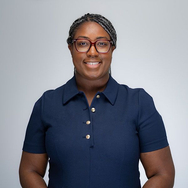 Bria Felton with glasses in navy blue shirt with button detail, grey background. Professional portrait.
