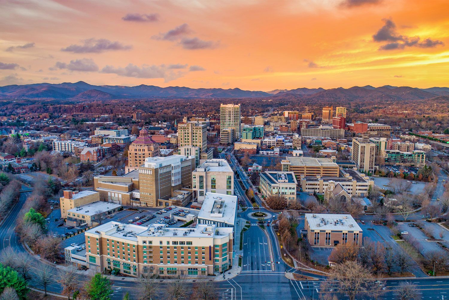Aerial view of Asheville cityscape at sunset, with mountains in the background and colorful sky.