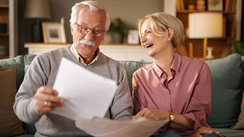 Elderly couple happily reviewing documents on a sofa at home, smiling and enjoying each other's company.