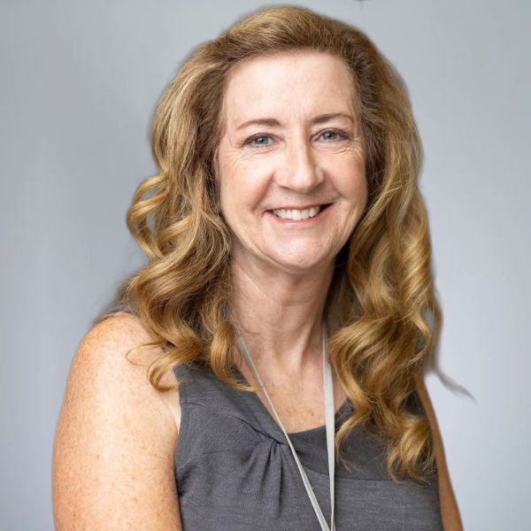 Mary Ellen McCoy with curly hair wearing a gray top, gray background, professional look.