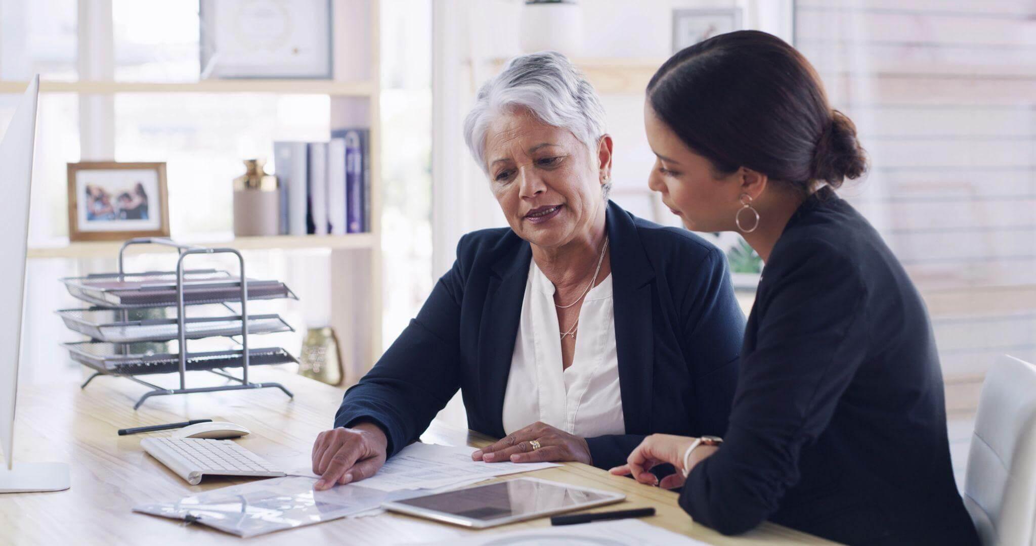 two women sitting at a desk looking at a tablet